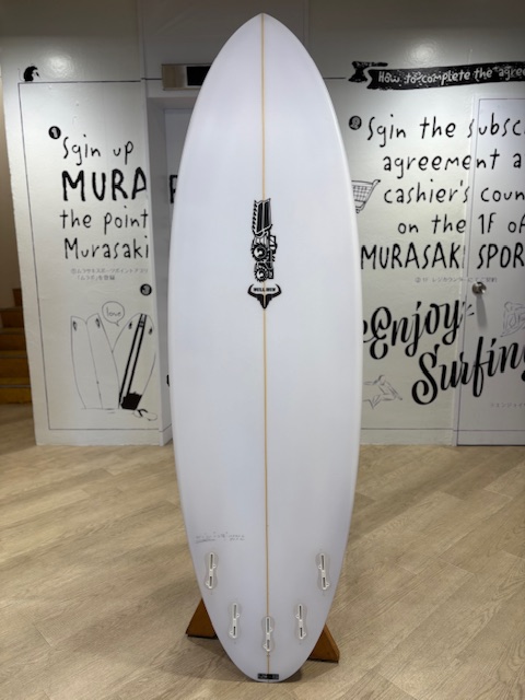 White surfboard on a wooden stand indoors, featuring a black emblem down the center and a thin yellow stringer, with graffiti-covered walls in the background.