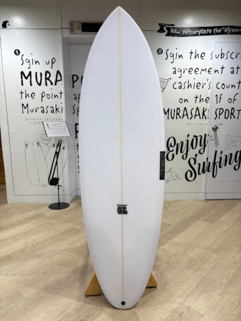 White surfboard on a wooden display stand in a showroom, centered against a wall with black handwritten text and drawings in the background.