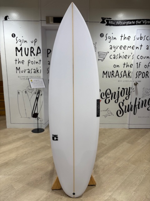 White surfboard standing upright on a wooden rack inside a gallery or showroom, with handwritten wall art in the background.