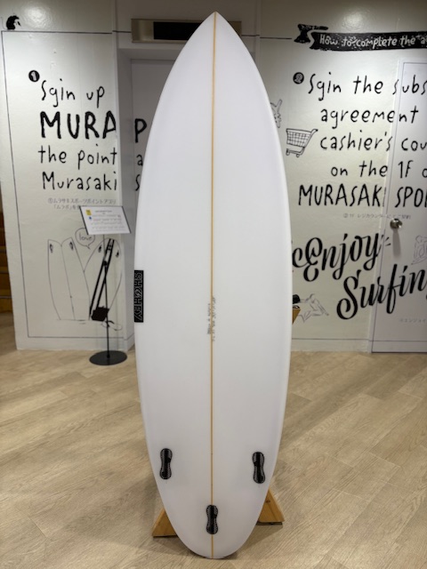 White surfboard with a thin yellow stringer, mounted upright on a wooden display stand in a gallery, with wall texts in the background.