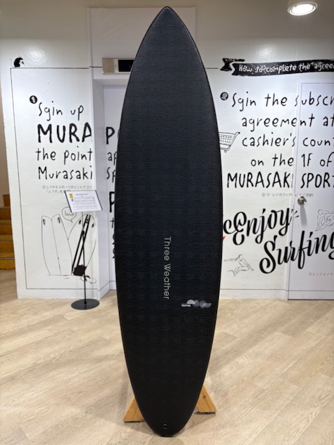 Black longboard surfboard on a wooden stand inside a showroom, with surfing-themed white walls; center of board labeled 'Three Weather'