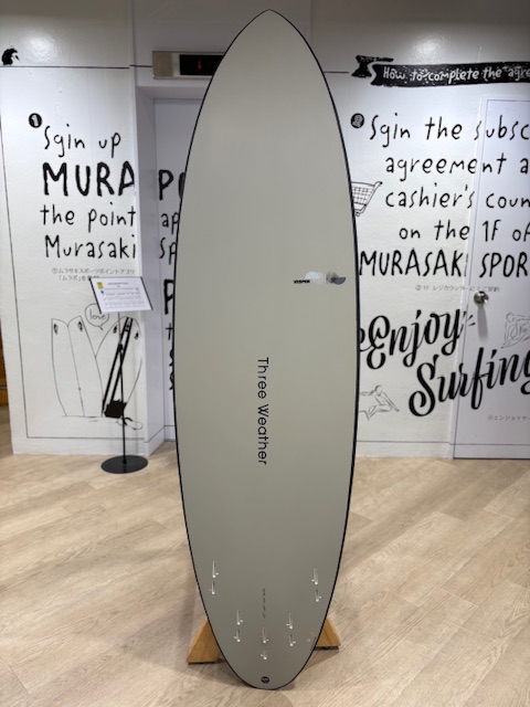 Gray surfboard on a wooden stand in a shop, with 'Three Weather' branding along the center