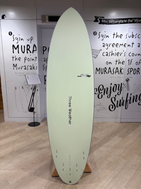 Mint-green surfboard on a wooden stand in a showroom; 'Three Weather' printed vertically with small logos near the top.
