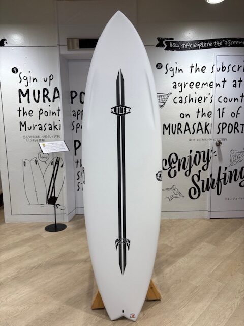 White surfboard with a black vertical stripe and Placebo logo, displayed upright on a wooden stand in a shop with handwritten wall art.