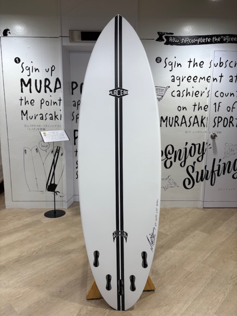 White surfboard with a black center stripe and logo, upright on a stand, showing four fin boxes near the bottom in a shop with graffiti-style walls.