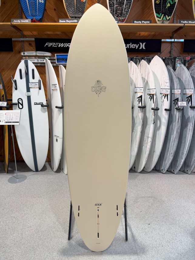 Beige surfboard on a stand in a surf shop, surrounded by other boards on racks.