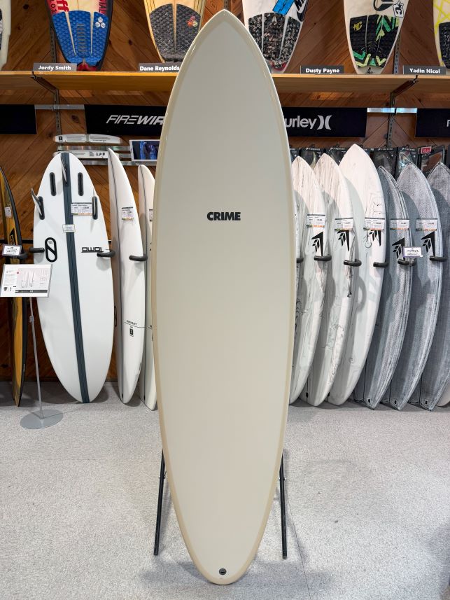 Beige longboard surfboard on a stand in a surf shop, brand CRIME visible, with other boards in the background