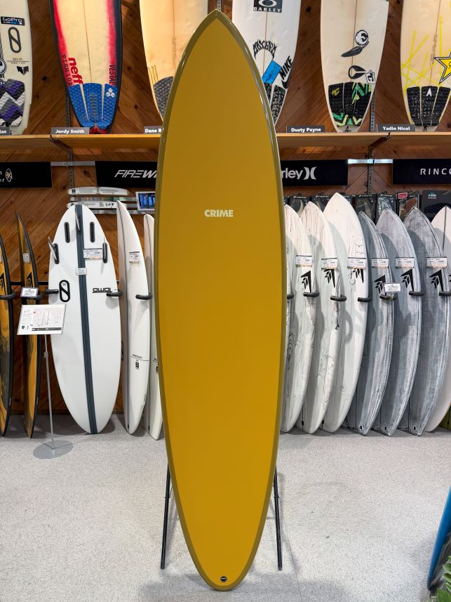 Yellow surfboard on a stand in a shop, with 'CRIME' logo near the center and other boards on racks behind it