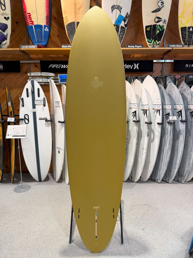 Olive-green surfboard on a display stand in a surf shop, with other boards on racks behind it.