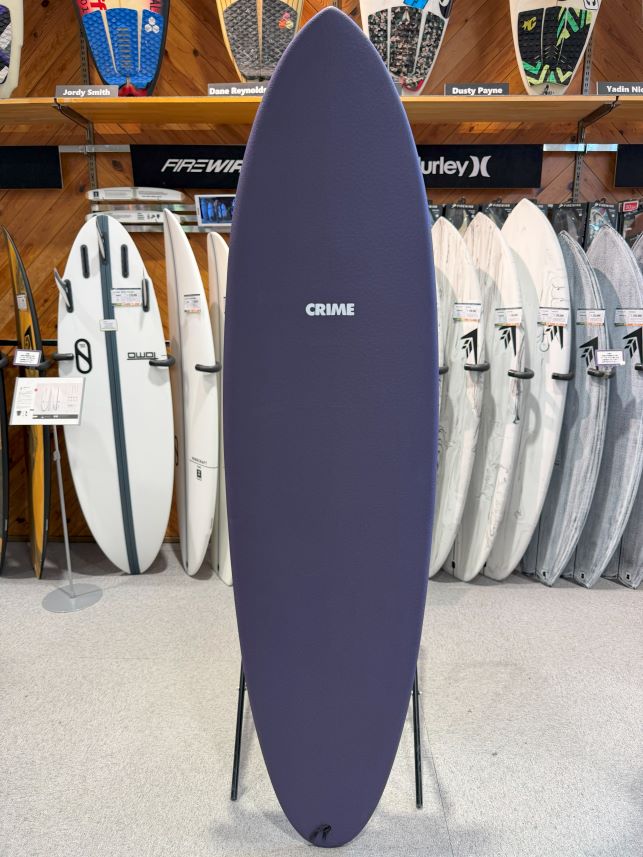 Tall purple surfboard on a stand in a surf shop, with racks of boards behind it labeled with brand names.