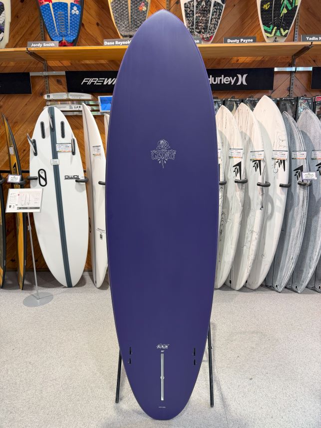 Purple surfboard on a display rack in a surf shop, surrounded by other boards on racks and shelves behind.