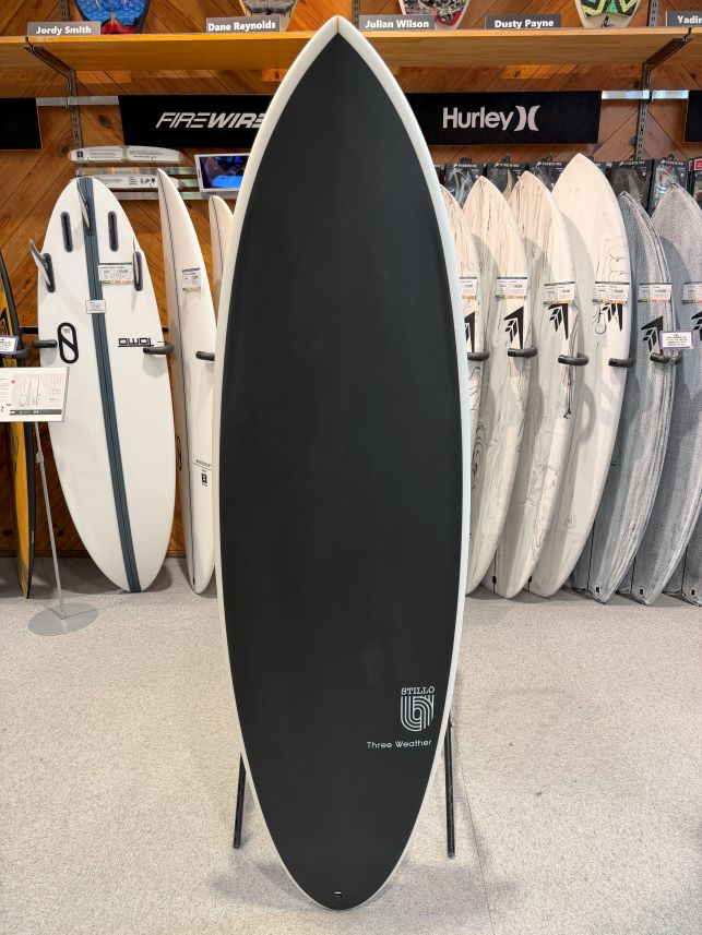 IMG_8602 - SHOP NEWS | ムラサキスポーツオンラインストア 通販 Black surfboard with white rails on a display stand in a surf shop, surrounded by white and gray boards in the background.