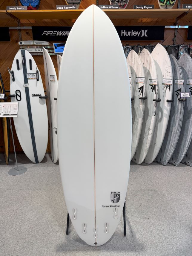 IMG_8606 - SHOP NEWS | ムラサキスポーツオンラインストア 通販 Large white surfboard on a display stand in a shop, surrounded by other boards and brand banners, including Hurley, in the background.