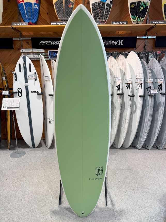 IMG_8608 - SHOP NEWS | ムラサキスポーツオンラインストア 通販 Green surfboard on a display stand in a surf shop, with white and gray boards on racks behind it.