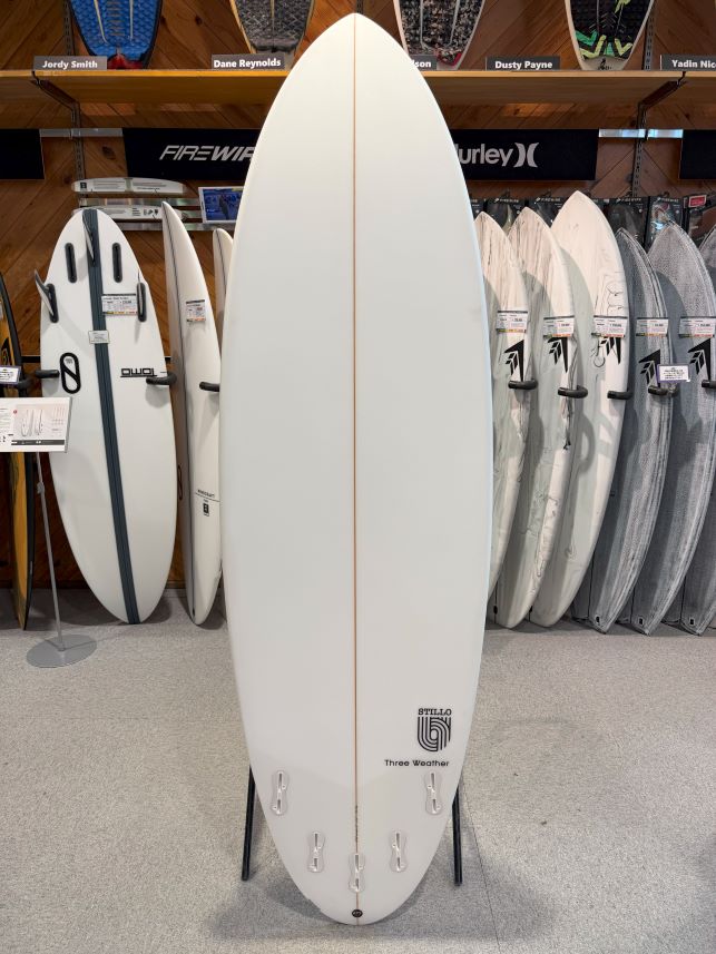 IMG_8609 - SHOP NEWS | ムラサキスポーツオンラインストア 通販 White surfboard on a display rack in a surf shop, standing upright with a light brown stringer and Three Weather logo near the tail, surrounded by other boards on shelves.