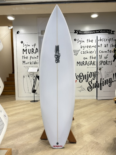 White surfboard on a wooden stand in a surf shop, featuring a small black graphic midboard and a red logo near the tail.