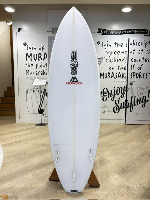White surfboard on a wooden stand indoors, with a MONSTA logo midboard and two fin boxes near the tail.