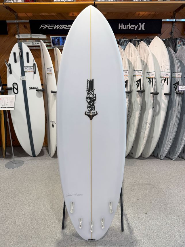 White surfboard on a stand in a surf shop, centered between other boards, featuring a vertical gold stringer and a bold logo near the middle.