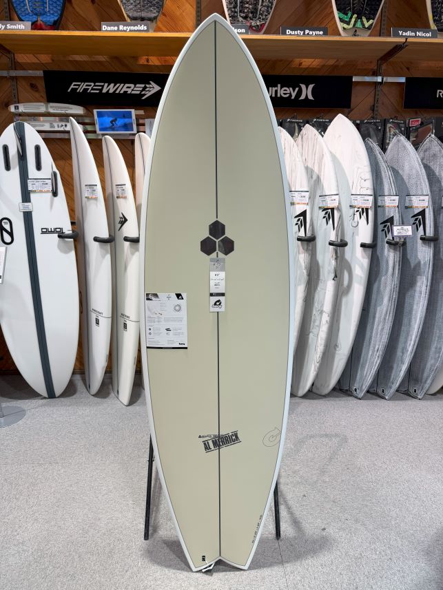Cream-colored surfboard on a stand in a shop, centered with a hexagon logo and price tag, with rows of other boards on racks in the background.