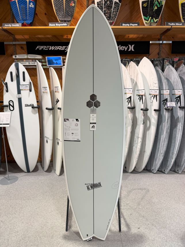 Tall light gray surfboard on a stand in a surf shop, surrounded by other boards on racks.