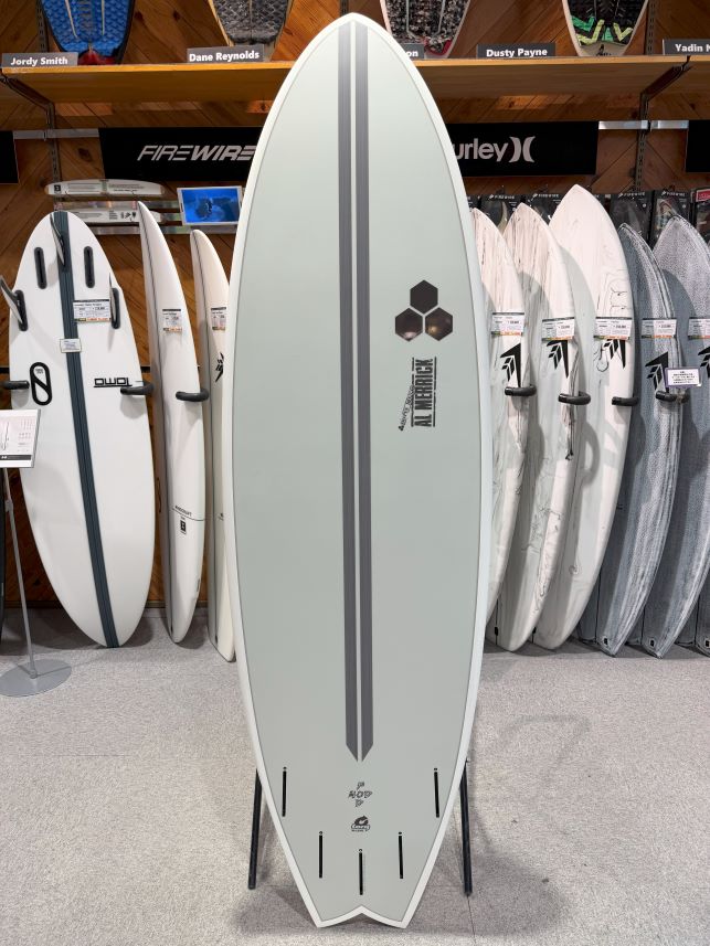 Center gray surfboard with a dark vertical stripe on a display stand in a surf shop, surrounded by other boards