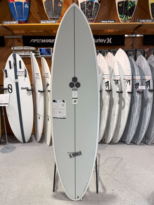 Gray surfboard on a display stand in a surf shop, surrounded by white and gray boards and wooden shelves with brand logos visible.
