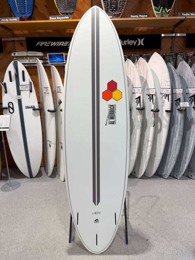 White surfboard with a centered gray double stripe and hex honeycomb logo (orange/red/yellow) near the upper middle, standing on a rack among other boards in a shop