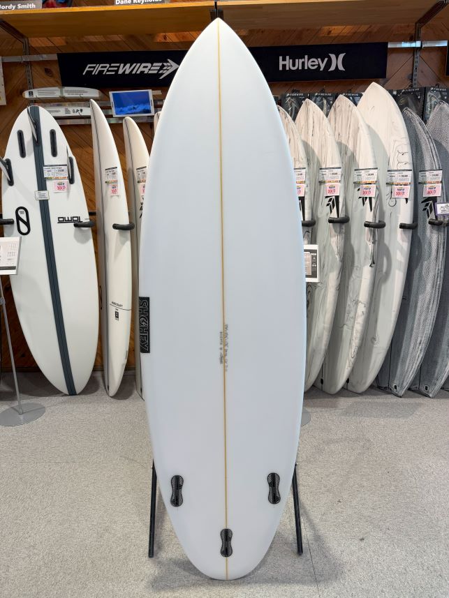 White surfboard standing upright on display stands in a surf shop, with a gold stringer down the center and four fin boxes at the tail. More boards line the background.