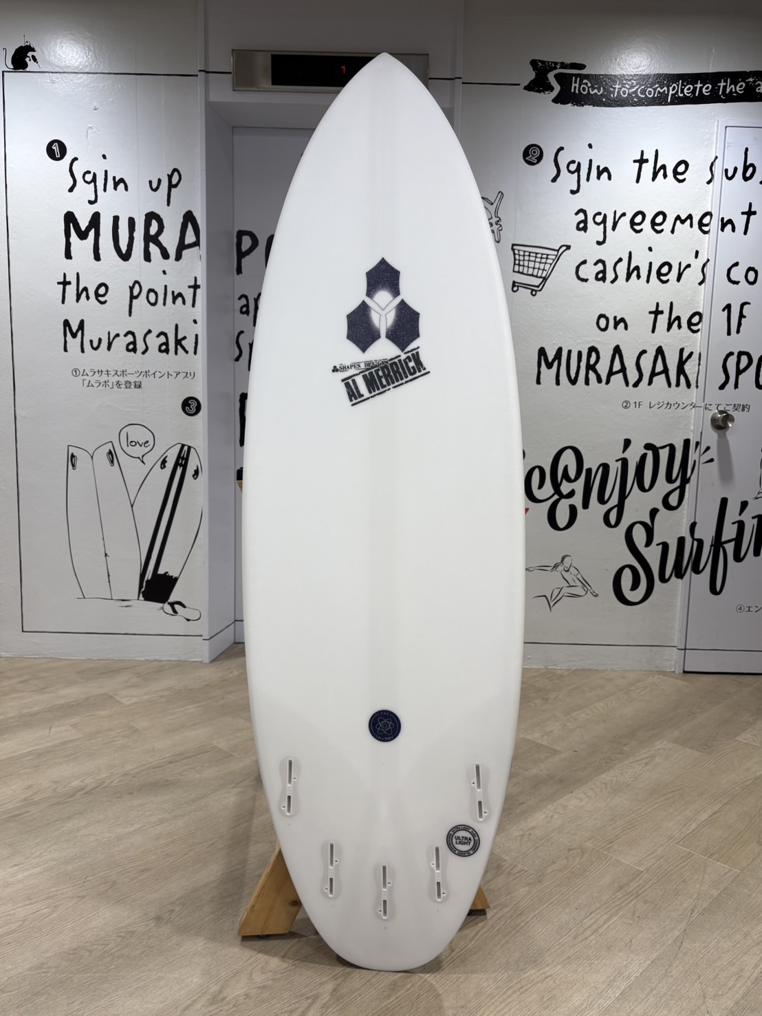 White surfboard on a wooden stand indoors with a blue hexagon logo and 'AL MERRICK' text on the chest area.