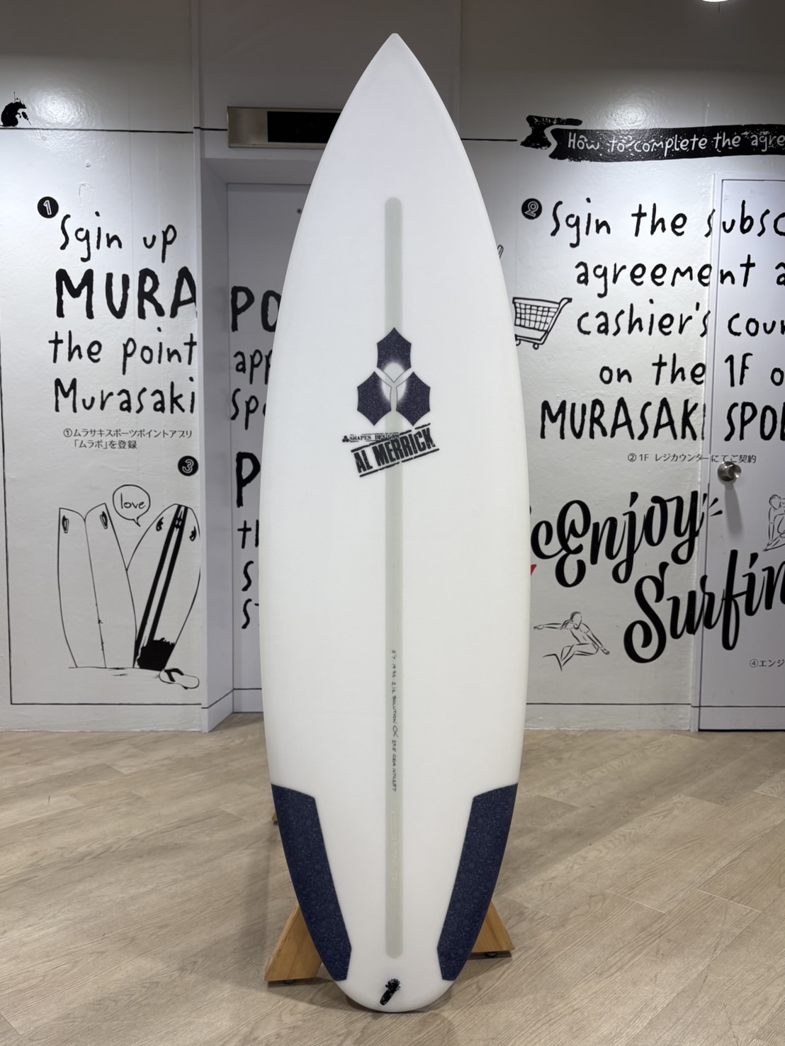 White surfboard on a wooden stand, standing upright indoors; features an Al Merrick logo midboard and dark blue tail pads/edges.