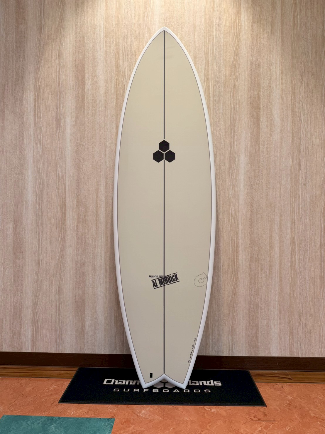 Beige surfboard standing upright against a wood-paneled wall on a display mat, featuring a center black stringer and hexagon logo near the middle.