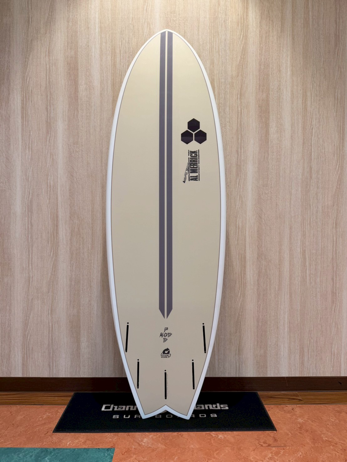 Beige surfboard with two dark vertical stripes and small logos, standing upright on a display mat against a wood panel wall.