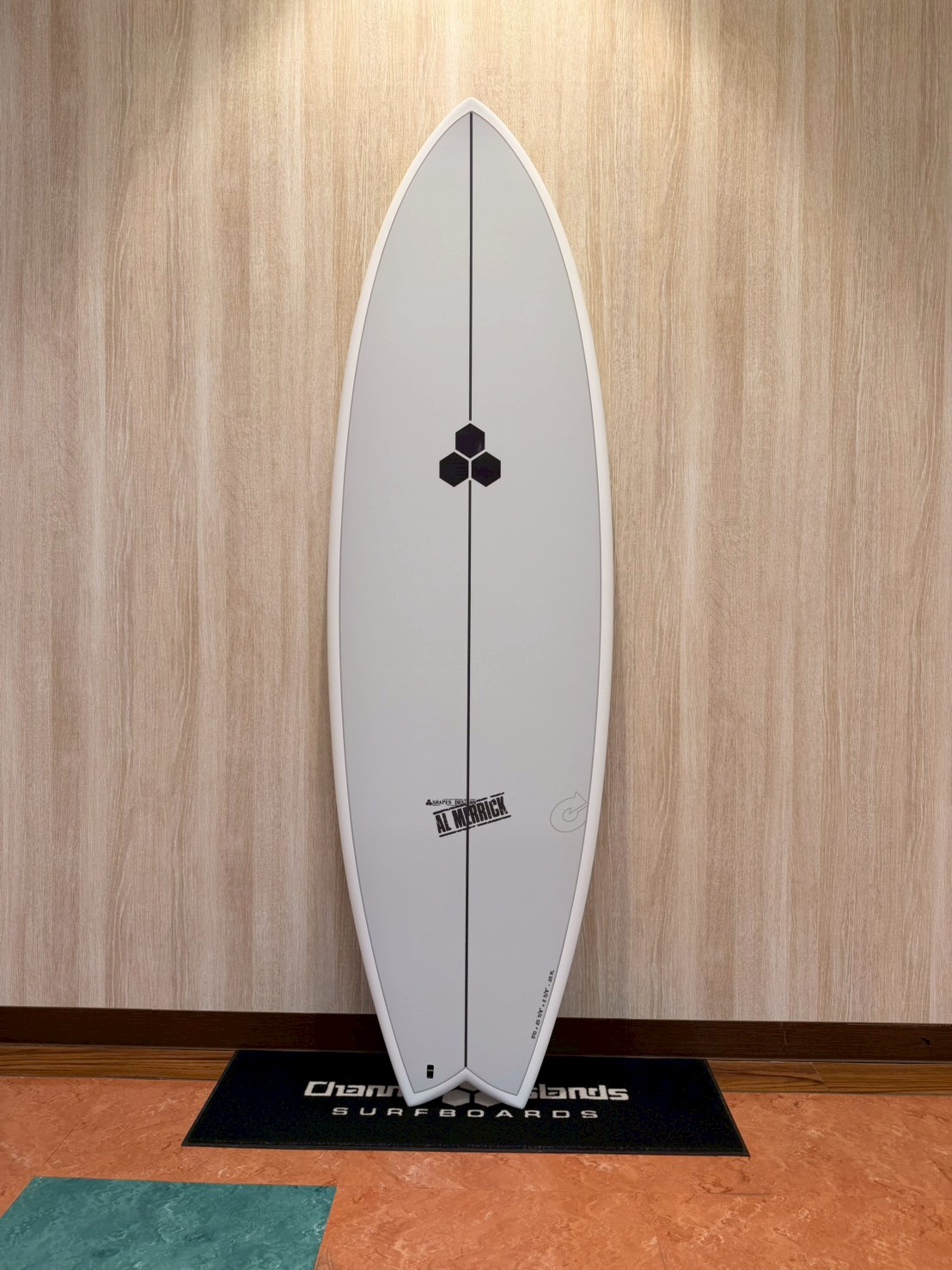 White surfboard on a display stand leaned against a wooden wall, with a black hexagon logo in the center and a vertical stringer, labeled Al Merrick at the bottom edge.