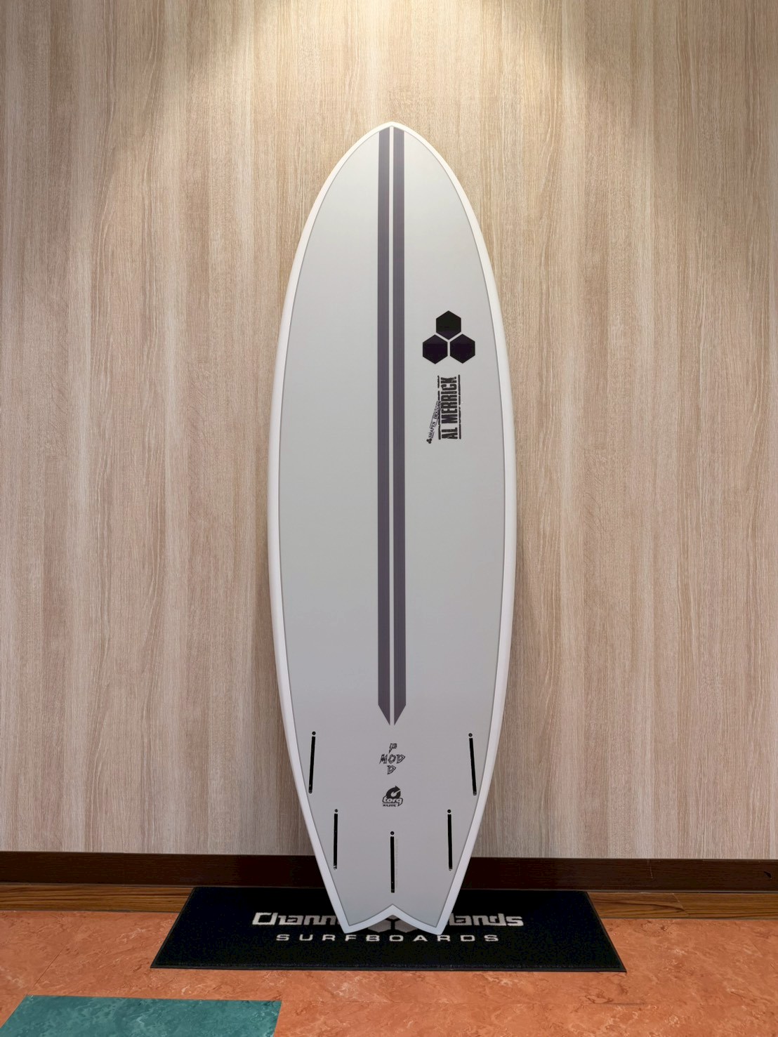 White surfboard standing upright against a wooden wall with a dark gray center stripe and logo near the middle, displayed indoors on a mat.