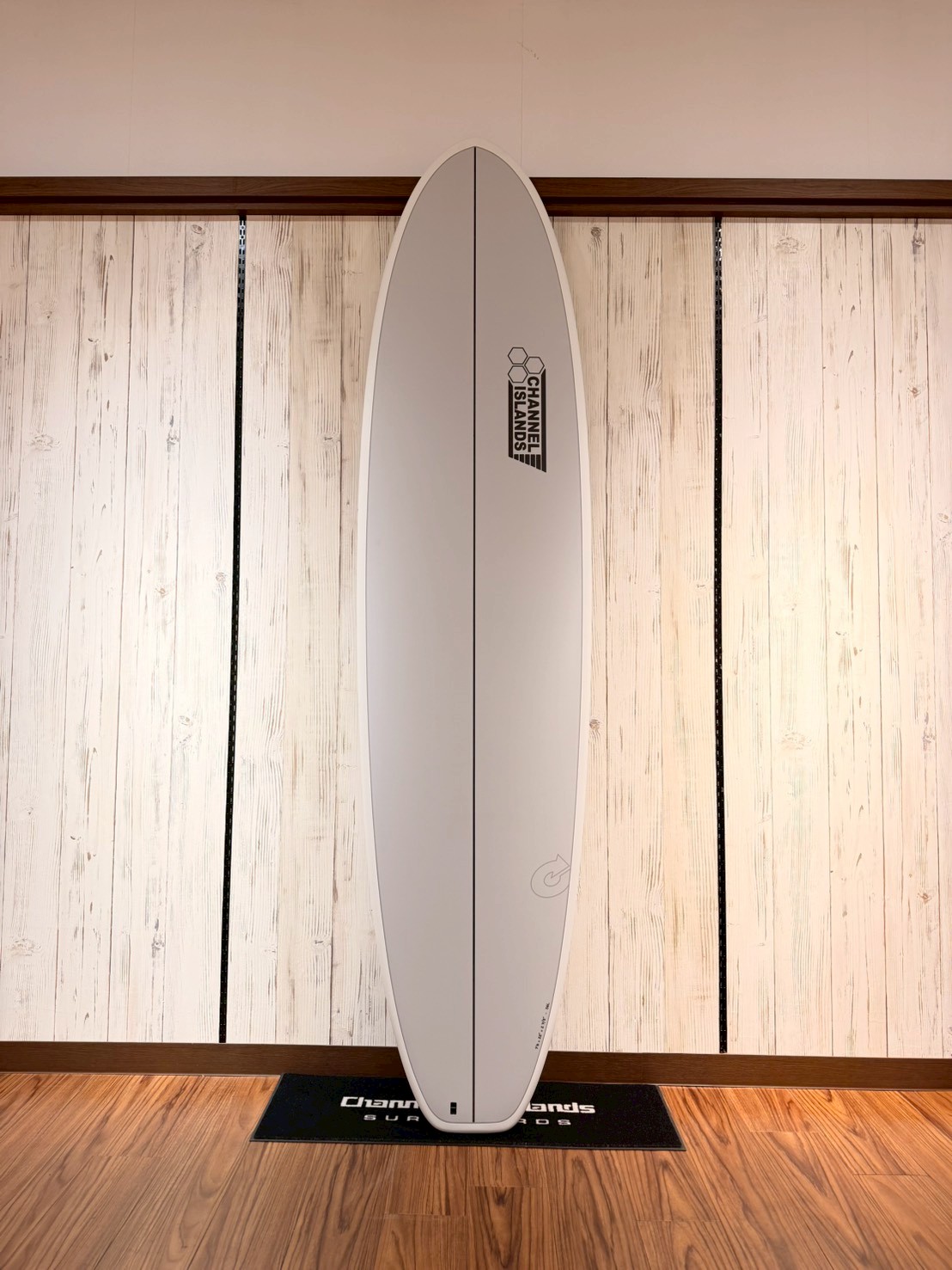 White surfboard standing upright against a light wood-paneled wall, centered on a black Channel Islands mat.