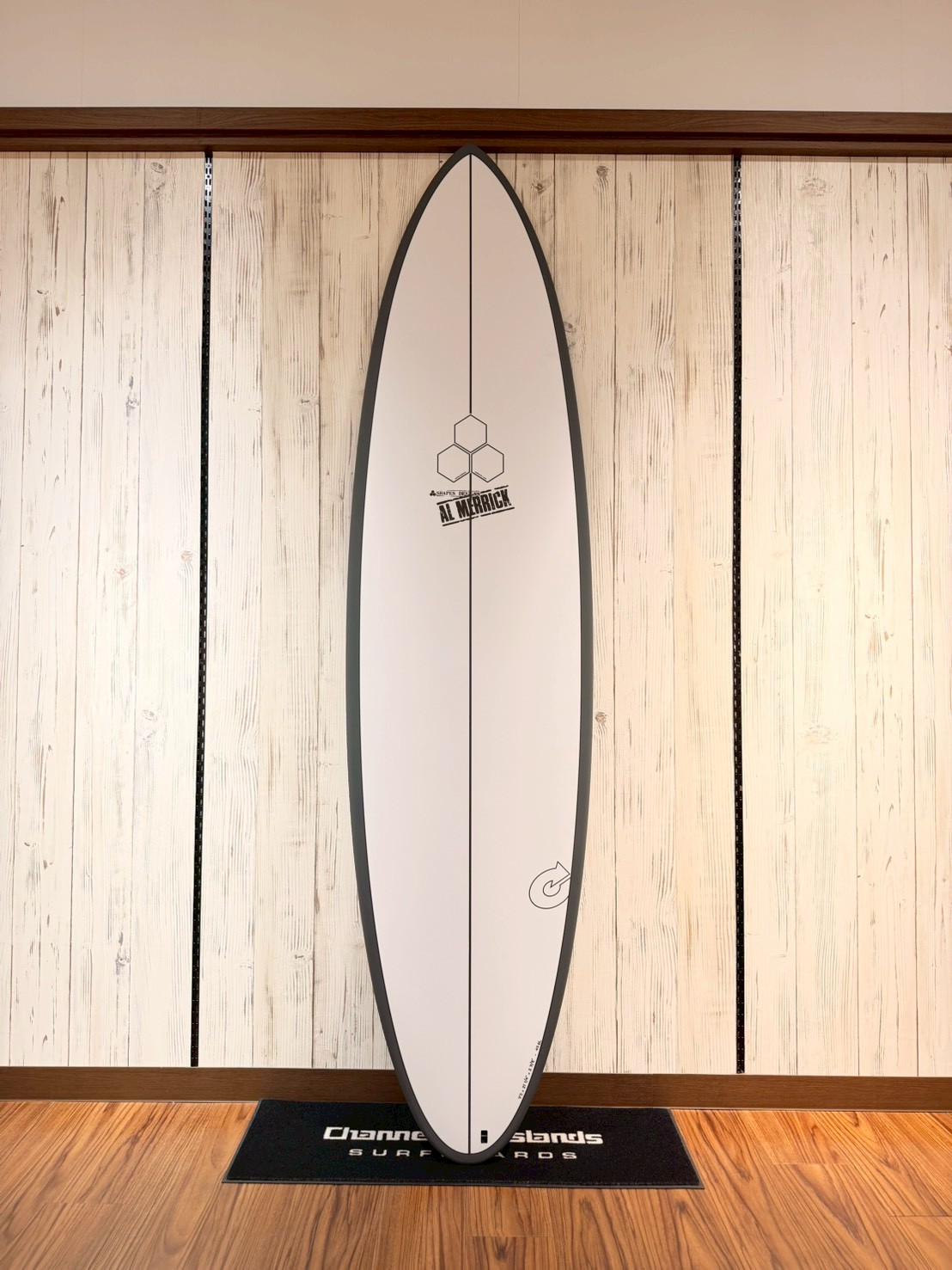 White surfboard with gray rails, standing upright on a wall display in a store, showing a hexagon logo and AL MERRICK text near the center.
