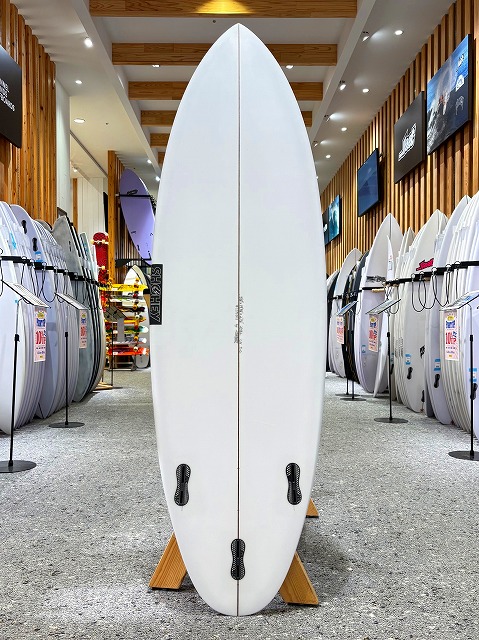 a-image0 - SHOP NEWS | ムラサキスポーツオンラインストア 通販 White surfboard on a display rack in a surf shop, centered with other boards lining the aisle behind it.