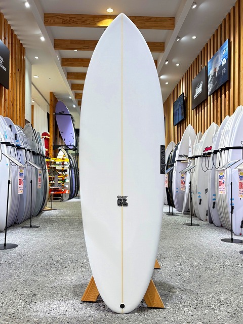 a-image1 - SHOP NEWS | ムラサキスポーツオンラインストア 通販 White longboard standing upright on a wooden rack inside a surf shop, with many other boards lining the walls.
