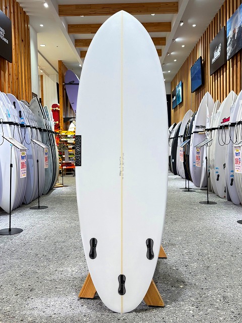 a-image2 - SHOP NEWS | ムラサキスポーツオンラインストア 通販 White surfboard standing upright on a wooden display stand in a surf shop, flanked by rows of boards on racks.