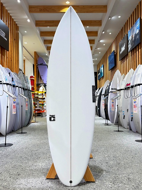 a-image3 - SHOP NEWS | ムラサキスポーツオンラインストア 通販 White surfboard standing upright on a wooden rack in a surf shop, with rows of boards lining the walls on both sides.