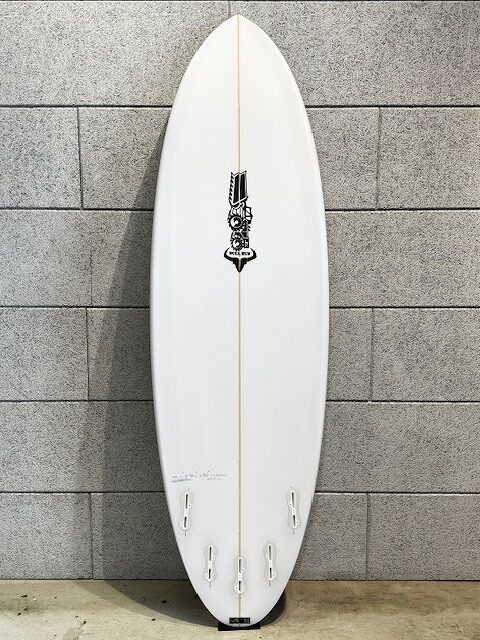 White surfboard with a center stringer leaning against a gray brick wall; features a black logo around the middle and three fin boxes at the tail.