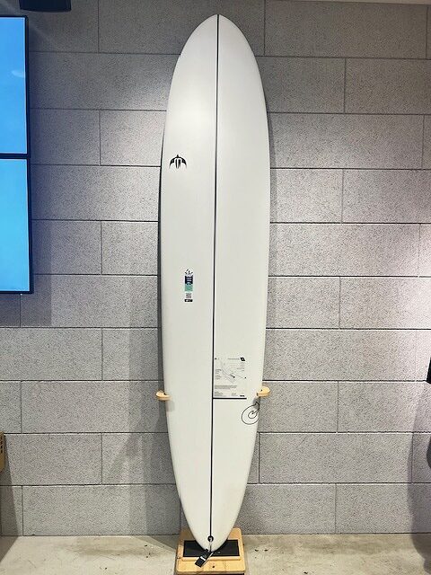 White surfboard standing upright on a wooden rack indoors against a gray tiled wall; longboard with subtle logos.
