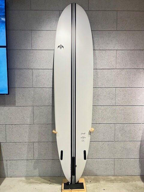 White surfboard with a double black center stripe standing upright on a wooden stand against a gray tiled wall, displayed indoors.