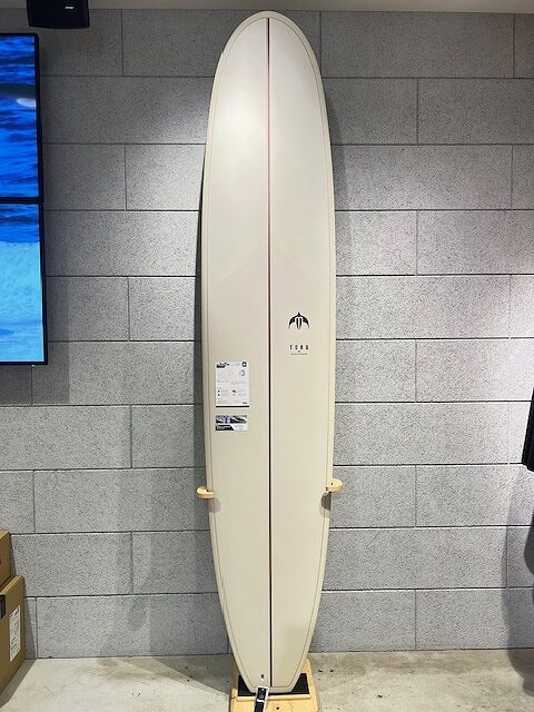 Cream-colored surfboard on a wooden stand against a gray tile wall, shown full length for display or sale.