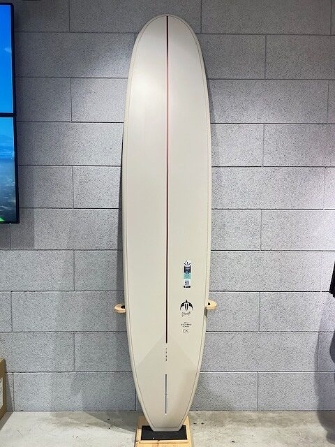 Cream surfboard with a red center stringer stands upright against a gray brick wall on a display rack in a store/ showroom setting.
