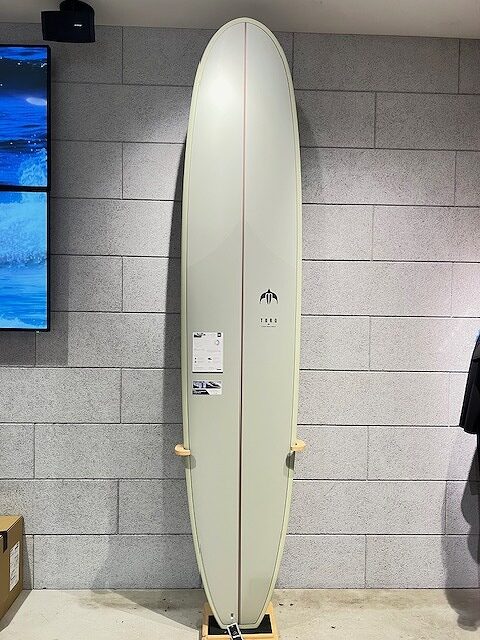 Cream-colored surfboard standing upright on a wooden rack indoors against a gray brick wall, with a small label visible on its center.