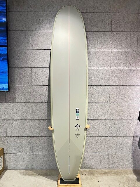 Cream surfboard standing upright on a wooden rack against a gray brick wall, features a red stringer and small logos midboard (indoor display).