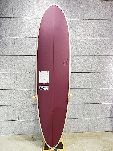 Maroon longboard surfboard on a display stand, upright against a gray brick wall; small stickers on the deck.
