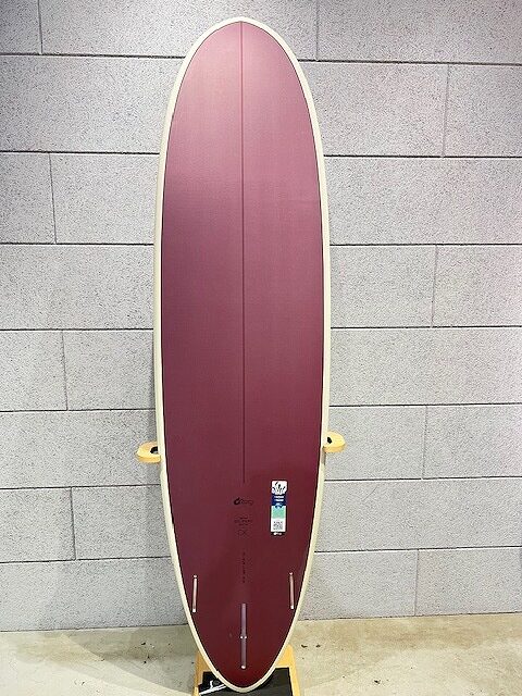 Maroon longboard with cream rails standing upright on a wooden rack against a gray brick wall; yellow trucks visible on the sides and a sticker near the center.