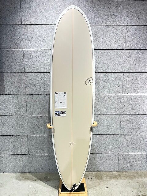 Cream-colored surfboard standing upright on a wooden stand against a gray tiled wall, with labels on the deck.